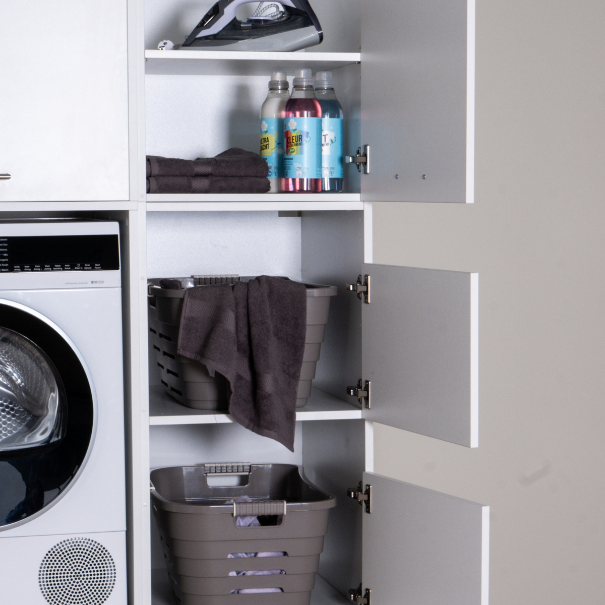 White laundry cabinet with open doors, storage shelves holding folded towels, soda bottles, and two brown laundry baskets.